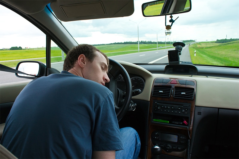 man falling asleep behind the wheel of his car on the side of the road